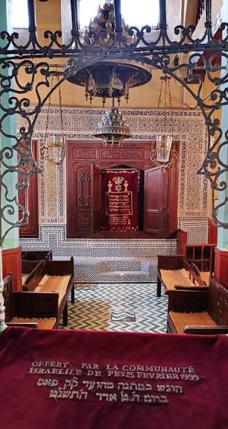 The interior of the Ibn Danan Synagogue in the Fes Mellah viewed through the ornate wrought iron entrance gate — the bimah and Torah ark are visible beyond, framed by zellige tilework and carved plaster in the Andalusian-Moroccan tradition